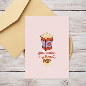 May include: A pink card with a cartoon illustration of a red and white popcorn box with a heart on it. The text on the card reads "You make my heart POP".