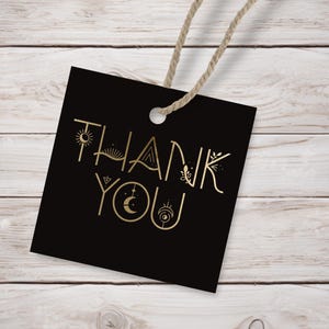 Könnte beinhalten: Schwarzes Dankeschön-Etikett mit goldener Aufschrift "Danke" und dekorativen Elementen um die Buchstaben. Das Etikett ist an einem Stück Schnur befestigt.
