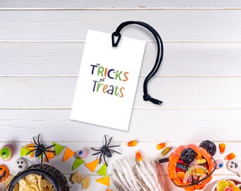 Może przedstawiać: Biała etykieta z napisem "No Tricks Just Treats" w kolorze zielonym, pomarańczowym i niebieskim wisi na czarnym sznurku. Dekoracje o tematyce Halloween, w tym pająki, cukierki i wiadro z dyni, otaczają etykietę na białej drewnianej powierzchni.