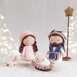 Może przedstawiać: Szydełkowa szopka bożonarodzeniowa z Maryją, Józefem, dzieciątkiem Jezus w żłóbku, barankiem i gwiazdą na patyku. Figurki są głównie beżowe, różowe i brązowe, z niebieskimi i złotymi akcentami. Tło jest miękkie i rozmyte.