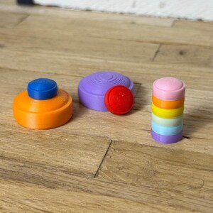Könnte beinhalten: Eine Auswahl an farbenfrohen, 3D-gedruckten Objekten. Es gibt ein orangefarbenes und blaues Teil, eine lila Scheibe mit einem roten Kreis und einen Stapel regenbogenfarbener Zylinder. Die Artikel sind auf einer Holzoberfläche angeordnet.