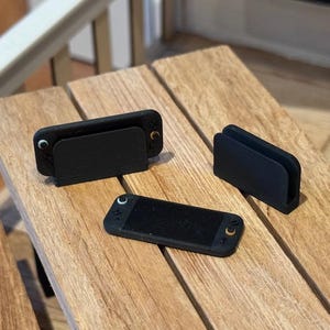 May include: Three black handheld gaming devices and accessories are arranged on a wooden picnic table. The table has a natural wood grain finish. The devices are rectangular with rounded edges and small orange accents.