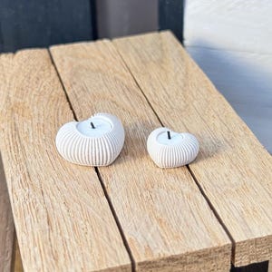 Op de afbeelding: Twee witte hartvormige kandelaars met een gestructureerd, geribbeld ontwerp staan op een lichtbruin houten oppervlak. De kaarsen zijn niet aangestoken. Het houten oppervlak maakt deel uit van een kleine tafel.