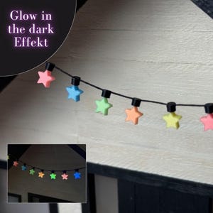 Può includere: Una stringa di luci decorative a forma di stella in rosa, blu, verde, arancione e giallo. Le luci sono infilate su un filo nero e montate su un muro bianco. Il testo "Glow in the dark Effekt" è visibile.