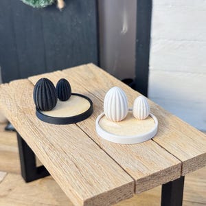 Könnte beinhalten: Zwei Sätze dekorativer Eierskulpturen auf einem Holztisch. Ein Satz zeigt schwarze Eier auf schwarzem Sockel, der andere weiße Eier auf weißem Sockel. Der Tisch hat eine natürliche Holzoberfläche.