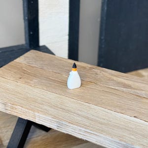 May include: A miniature white glue bottle with a black and orange cap sits on a small wooden table. The table has a light brown finish and black legs. The background includes a window and a black and white wall.