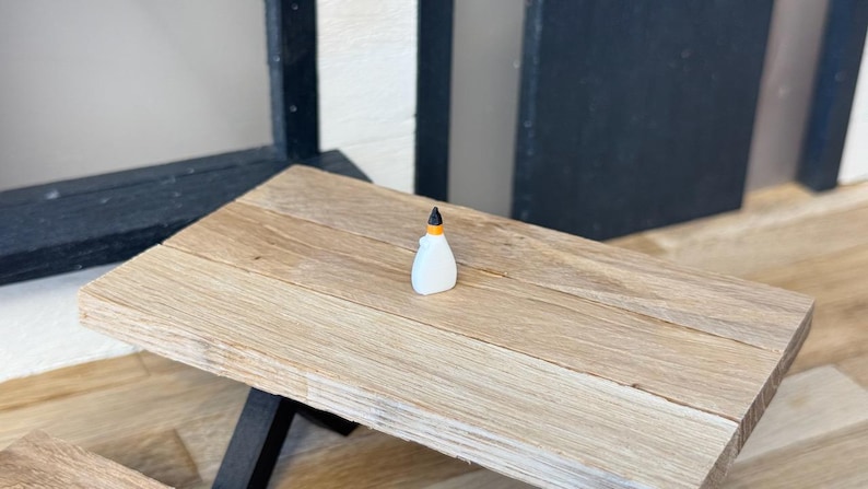 May include: A miniature white glue bottle with a black and orange cap sits on a small wooden table. The table has a light brown finish and black legs. The background includes a window and a black and white wall.