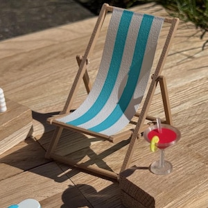 May include: Miniature wooden beach chair with blue and white striped fabric. A small cocktail with a red liquid and a lime slice sits nearby. A bottle of sunscreen and other beach accessories are also present.