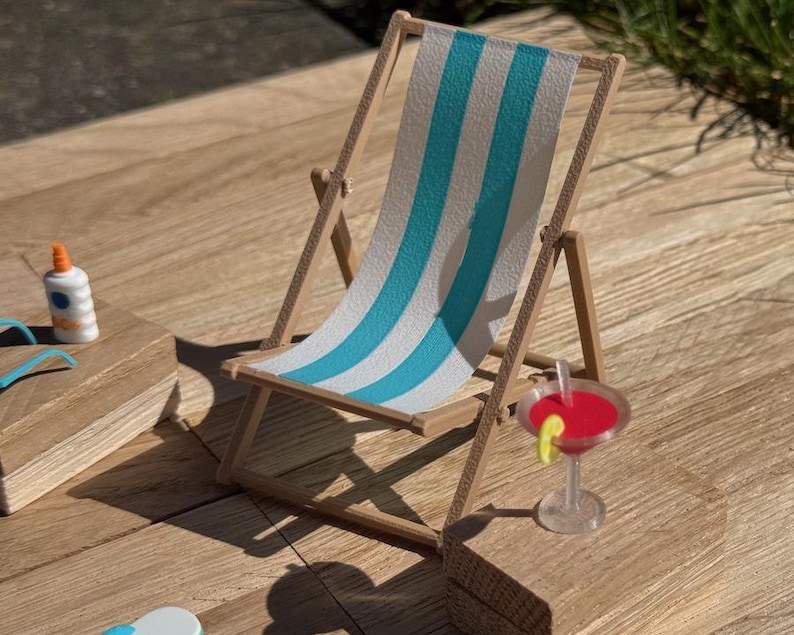 May include: Miniature wooden beach chair with blue and white striped fabric. A small cocktail with a red liquid and a lime slice sits nearby. A bottle of sunscreen and other beach accessories are also present.