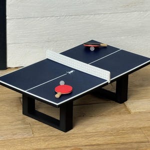 Könnte beinhalten: Miniatur-Tischtennistisch in Marineblau mit weißem Netz, weißen Linien und schwarzen Beinen. Enthält zwei Schläger, einen roten und einen schwarzen, und zwei weiße Tischtennisbälle. Ein lustiges, dekoratives Objekt.