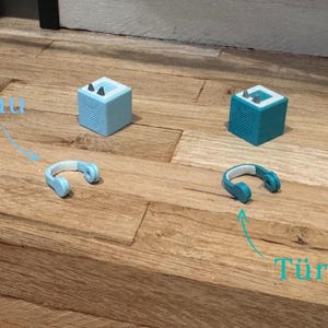 May include: Two small, cube-shaped speakers, one light blue and one turquoise, with headphones in the same colors. The light blue speaker is labeled "Blau" and the turquoise speaker is labeled "T&uuml;rkis".