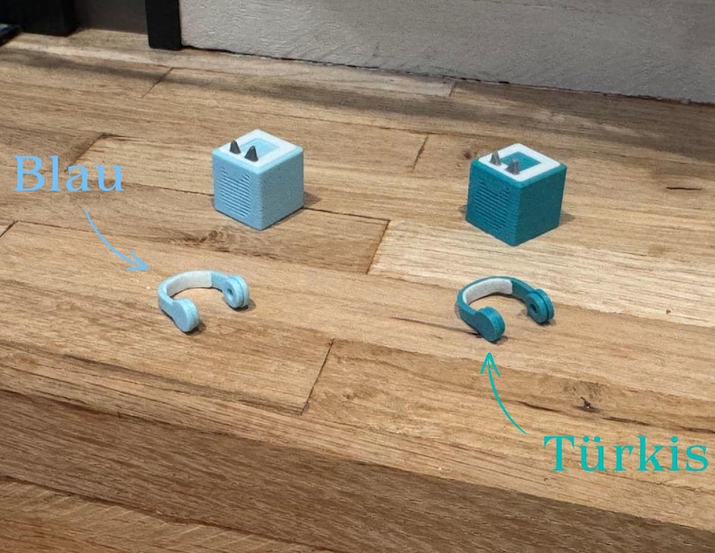 May include: Two small, cube-shaped speakers, one light blue and one turquoise, with headphones in the same colors. The light blue speaker is labeled "Blau" and the turquoise speaker is labeled "T&uuml;rkis".