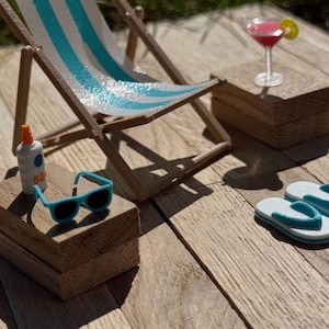 May include: A miniature beach scene with a striped deck chair, sunglasses, sunscreen, a cocktail, and flip-flops. The chair has blue and white stripes. The sunglasses are turquoise. The cocktail has a lemon slice. The scene is set on a wooden surface.