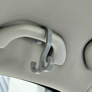 Può includere: Un gancio in plastica grigia progettato per appendere oggetti alla maniglia superiore di un'auto. Il gancio è fissato alla maniglia con un semplice clip.