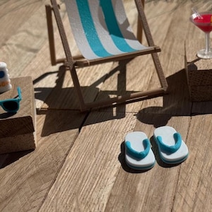 May include: Miniature beach scene with a striped blue and white beach chair, sunglasses, sunscreen, a cocktail, and flip-flops. The scene is set on a wooden surface, evoking a sunny day at the beach.