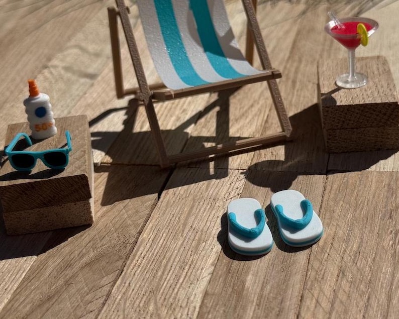 May include: Miniature beach scene with a striped blue and white beach chair, sunglasses, sunscreen, a cocktail, and flip-flops. The scene is set on a wooden surface, evoking a sunny day at the beach.