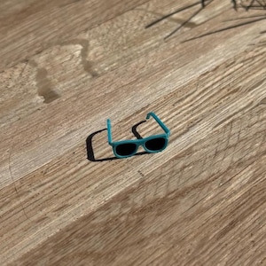 May include: Small, turquoise sunglasses with black lenses rest on a wooden surface. The sunglasses have a classic rectangular shape and a bright, solid color frame. The image is taken in natural light.