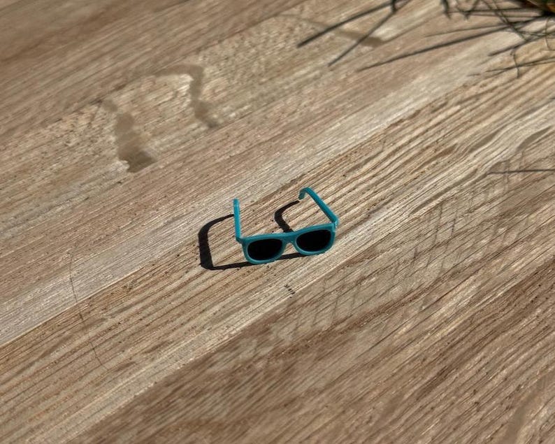 May include: Small, turquoise sunglasses with black lenses rest on a wooden surface. The sunglasses have a classic rectangular shape and a bright, solid color frame. The image is taken in natural light.
