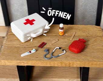 Op de afbeelding: Een witte miniatuur eerstehulpkoffer met een rood kruis en de tekst "ÖFFNEN" met een pijl, vergezeld van een klein medicijnflesje, een pleister, een houten stokje, een rode warmwaterkruik, een strip met rode pillen en een stethoscoop.