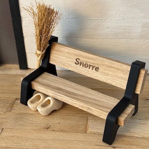 Könnte beinhalten: Eine Miniatur-Holzbank mit dem Namen "Snorre" auf dem Sitz eingraviert. Die Bank hat schwarze Stützen und hält einen kleinen Besen und ein Paar winzige Holzschuhe. Die Bank steht auf einer Holzoberfläche.