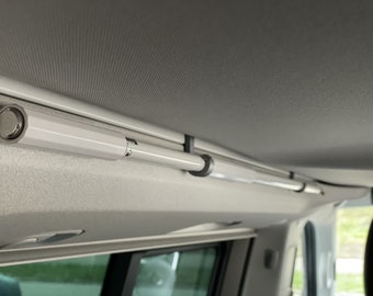 Op de afbeelding: Een witte intrekbare gordijnroede gemonteerd aan het plafond van een voertuig. De roede is uitgetrokken en wordt op zijn plaats gehouden door twee zwarte clips.