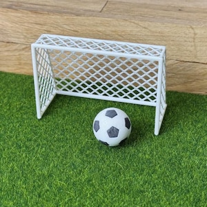 Könnte beinhalten: Ein weißes Miniatur-Fußballtor mit einem rautenförmigen Netzmuster, das hinter einem kleinen, runden Fußball mit klassischem schwarz-weißem Fünfeck-Design steht und auf einer strukturierten grünen Oberfläche platziert ist.