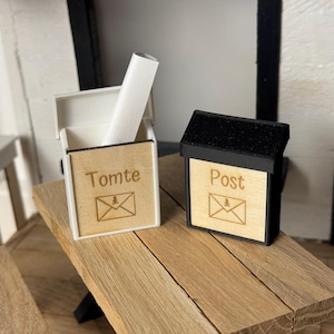 Op de afbeelding: Twee miniatuur houten brievenbussen, één witte en één zwarte, staan op een houten oppervlak. De witte brievenbus heeft het woord "Tomte" en een envelopafbeelding. De zwarte brievenbus heeft het woord "Post" en een envelopafbeelding.