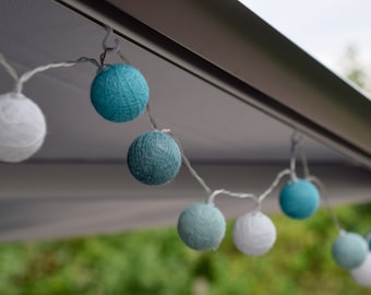 Op de afbeelding: Een string met witte en blauwe katoenen ballen die aan een wit plafond hangt. De lampjes zijn met een doorzichtige draad aan elkaar verbonden.