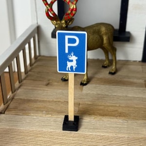 Könnte beinhalten: Ein blaues Miniatur-Parkschild mit weißem Rand und weißer Hirschgrafik. Das Schild ist auf einem Holzpfosten mit schwarzem Sockel montiert. Im Hintergrund befindet sich ein goldfarbenes Rentier mit roten und goldenen Geweihen.