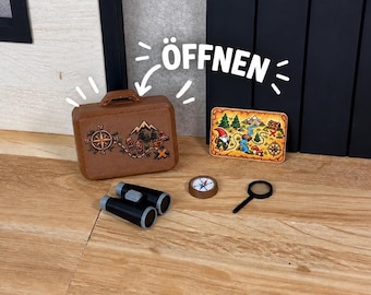 Könnte beinhalten: Ein brauner Miniaturkoffer mit Kompass- und Bergmotiv, eine rechteckige Schatzkarte mit einem Zwerg, ein schwarzes Fernglas, ein kleiner runder Kompass und eine schwarze Lupe. Der Text ÖFFNEN ist zu sehen.