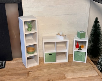 Peut inclure: Trois étagères cubiques blanches avec différents objets dessus. L'étagère de gauche a une figurine de lapin vert et un bol brun. L'étagère du milieu a une boîte verte et une boîte de mouchoirs blanche. L'étagère de droite a une décoration rouge et jaune, une décoration d'arbre vert et une boîte verte.