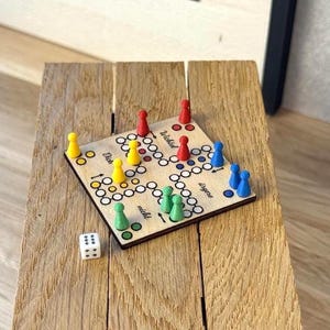 Könnte beinhalten: Ein quadratisches Brettspiel aus Holz mit einem Raster aus kreisförmigen Feldern und schwarzem Textaufdruck. Das Spielbrett enthält vier Sätze farbiger Spielfiguren in Rot, Gelb, Grün und Blau sowie einen weißen sechsseitigen Würfel mit schwarzen Punkten.