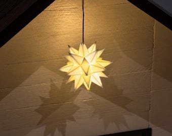 Può includere: Una lanterna di carta a forma di stella con molteplici punte, appesa a un sottile cordino nero. La lanterna è illuminata dall'interno, diffondendo una luce calda attraverso i pannelli di carta bianca e proiettando ombre geometriche.