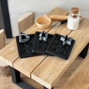 May include: Four black cookie cutters with different shapes, including a star, a tree, and a crown. The cutters are on a wooden table.