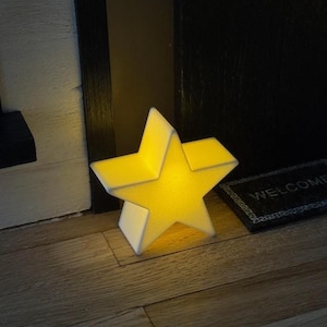 Op de afbeelding: Een oplichtende, stervormige decoratieve lamp met een witte basis en een gele, getextureerde voorkant. Het licht staat op een houten vloer naast een kleine welkomstmat. De ster is ongeveer 20 cm hoog.