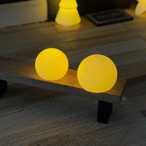 Op de afbeelding: Twee gloeiende gele bolvormige decoratieve lampen staan op een houten bankje met zwarte poten. De lampen hebben een textuur en geven een warme gloed. Andere decoratieve lampen zijn op de achtergrond.