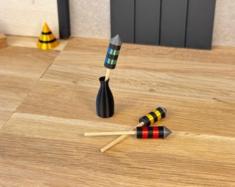 May include: Three miniature rocket-shaped decorations with wooden sticks and pointed tips. One rocket is teal and black, one is yellow and black, and one is red and black. They are displayed with a small black vase and other small items.