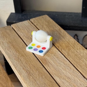 Op de afbeelding: Een klein wit eivormig object staat op een witte rechthoekige basis met kleurrijke stippen. De basis heeft gele accenten. Het object staat op een houten oppervlak.