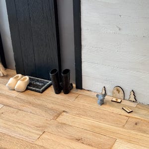 Op de afbeelding: Een miniatuur houten deur met een welkom bord, twee kleine bomen en een grijs muis figuur op een houten vloer. De deur is omgeven door witte houten panelen.