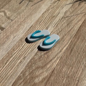 May include: Two miniature flip-flops. The sandals are gray with a blue strap and a white border. They are placed on a wooden surface with a natural wood grain pattern. The flip-flops are a miniature representation of summer footwear.