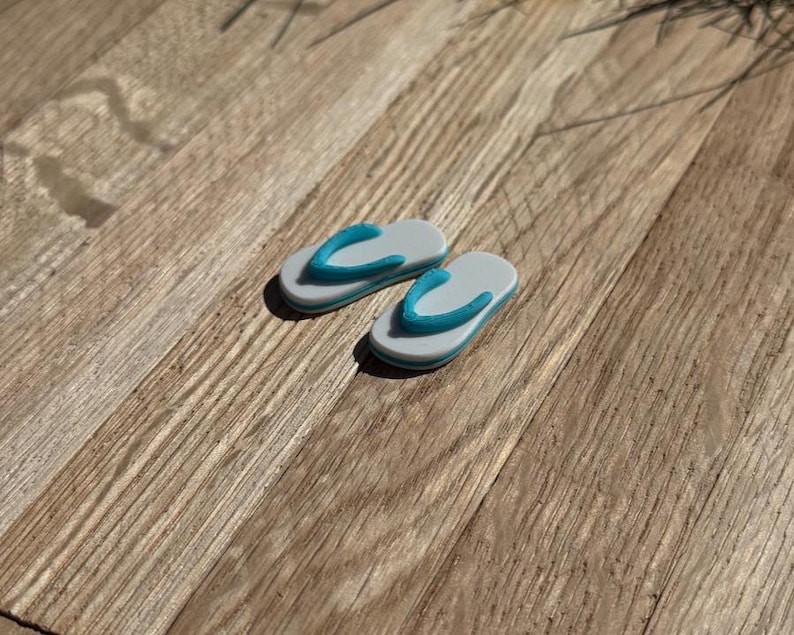 May include: Two miniature flip-flops. The sandals are gray with a blue strap and a white border. They are placed on a wooden surface with a natural wood grain pattern. The flip-flops are a miniature representation of summer footwear.