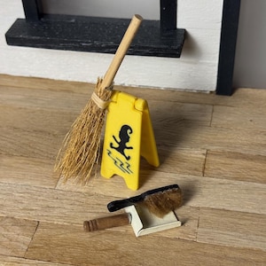 Könnte beinhalten: Miniatur-Reinigungsset mit Besen, Kehrschaufel und gelbem "Achtung Rutschgefahr"-Schild. Der Besen hat einen Holzgriff und Strohborsten. Die Kehrschaufel ist weiß mit schwarzem Griff und Bürste. Das Schild hat eine schwarze Silhouette.
