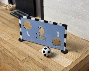 May include: A miniature soccer goal with a light blue backboard featuring a cutout of a gnome and two circular openings. A black and white soccer ball sits in front of the goal. The goal has black and white striped posts.
