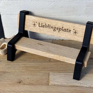 Könnte beinhalten: Eine Miniatur-Holzbank mit schwarzen Stützen und einer hellbraunen Sitzfläche und Rückenlehne. Die Rückenlehne ist mit dem Wort "Lieblingsplatz" graviert. Zwei kleine, weiße Holzschuhe befinden sich neben der Bank. Die Szene spielt auf einer Holzoberfläche.