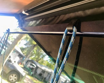 May include: A close-up of a black plastic clip attached to a blue rope with yellow and white stripes. The rope is attached to a black metal bar.