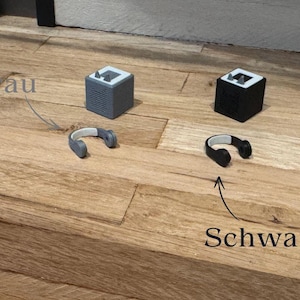 May include: Two small, cube-shaped speakers, one gray and one black, with matching headphones. The gray speaker has the word "Grau" written above it, and the black speaker has the word "Schwarz" written below it.