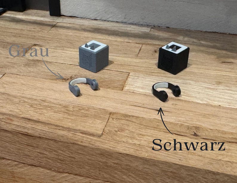 May include: Two small, cube-shaped speakers, one gray and one black, with matching headphones. The gray speaker has the word "Grau" written above it, and the black speaker has the word "Schwarz" written below it.