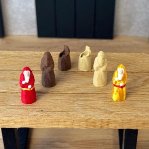 Op de afbeelding: Een verzameling van zes miniatuurbeeldjes in de vorm van figuren met capuchon en gewaad. De beeldjes zijn uitgevoerd in diverse effen kleuren, waaronder rood, donkerbruin, lichtbruin, beige en geel, elk met een klein wit gezichtsdetail.