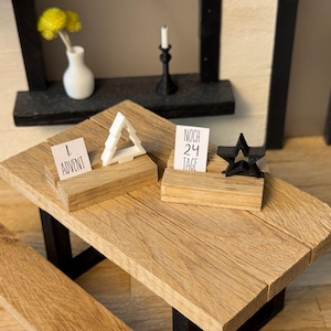Könnte beinhalten: Ein Miniatur-Holztisch mit zwei Holzblöcken, die Karten mit der Aufschrift "1. Advent" und "NOCH 24 TAGE" halten. Ein weißes Dreieck und ein schwarzer Stern befinden sich auf den Blöcken. Eine kleine Vase mit gelben Blumen und ein Kerzenhalter sind im Hintergrund.