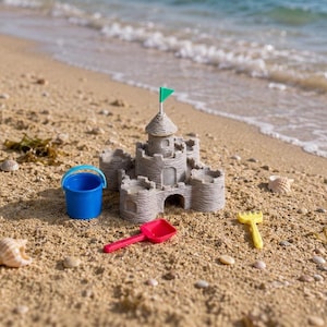 Pode incluir: Um castelo de areia de plástico bege com uma bandeira verde no topo, acompanhado por um balde azul brilhante com alça, uma pá de plástico vermelha e um ancinho de plástico amarelo, tudo disposto numa praia de areia perto da água do mar.